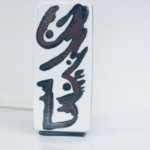 May include: White ceramic vase with a brown and black abstract design. The vase has a square shape and a wide mouth.