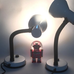 Op de afbeelding: Twee witte bureaulampen met flexibele zwanenhalsarmen en witte lampenkappen. De lampen zijn aan en verlichten een rood robot speelgoed in het midden van de afbeelding.