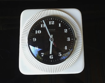 May include: White square kitchen timer with a black face and silver hands. The timer has a black rim with raised numbers and a black dial with the text "Kienzle International" in white.