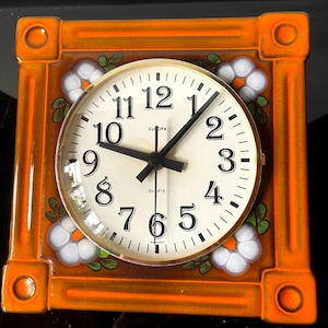 May include: Square orange quartz clock with a white face and black numerals. The clock face has black hands and the word "EUROPA" printed above the 12. The clock is decorated with white flowers and green leaves. The clock is framed in orange.