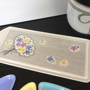 Könnte beinhalten: Ein weißes Keramik-Serviertablett mit einem grau-weißen Streifenmuster. Das Tablett zeigt ein Blumenmuster mit einem Strauß gelber, lila und blauer Blumen. Das Tablett hat zwei Griffe an den Seiten.