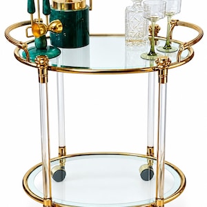 May include: A round two-tier bar cart featuring a gold metal frame, clear acrylic supports, and two glass shelves. The cart includes four wheels and holds a green ice bucket, a set of green bar tools, a clear glass decanter, and two green glasses.