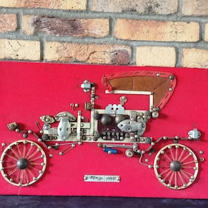 May include: A red felt board with a vintage car made of metal pieces, including screws, nuts, and wire. The car is labeled "Ford - 1903".