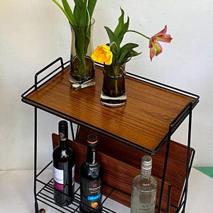 Könnte beinhalten: Ein schwarzer Barwagen aus Metall mit zwei Holzböden und einem Drahtgestell. Der Wagen hat vier goldene Räder und beherbergt drei Flaschen Alkohol und eine Vase mit gelben Tulpen.