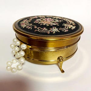Könnte beinhalten: Eine Vintage-Schmuckschatulle aus Messing mit einem schwarzen Samtdeckel mit floraler Stickerei in Rosa, Grün und Gelb. Die runde Schatulle hat einen Klappdeckel und steht auf drei kleinen, dekorativen Füßen. Eine Kette aus Kunstperlen liegt über der Seite.