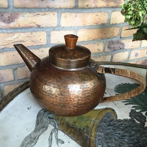 May include: A hammered copper teapot with a wooden knob on the lid and a woven rope handle.