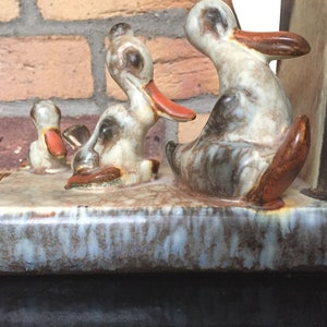 Könnte beinhalten: Ein Satz Keramik-Buchstützen mit drei Enten in einer Reihe. Die Enten sind in Braun-, Weiß- und Grautönen mit orangefarbenen Schnäbeln bemalt. Die Buchstützen stehen auf einem braun-weißen Sockel.