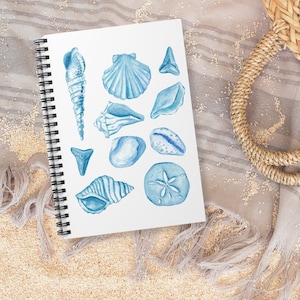 May include: A watercolor illustration of seashells and a starfish on a white page in a spiral bound sketchbook. The sketchbook is lying on a light brown sandy surface with a light gray fringed fabric in the background.