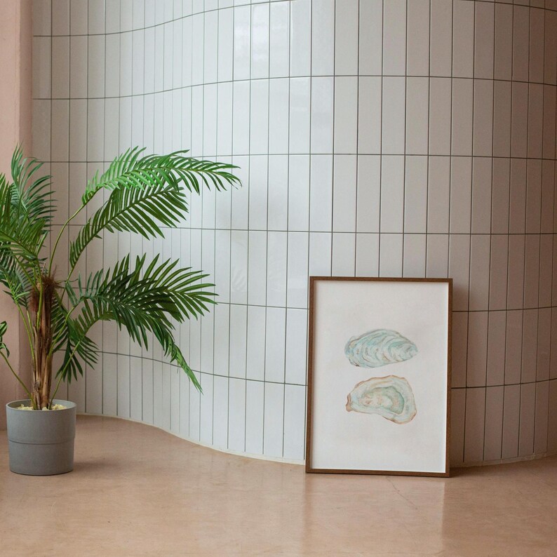 Puede incluir: Una impresi&oacute;n enmarcada de dos pinturas de acuarela de conchas de ostras en una pared de azulejos blancos con una palmera en maceta en primer plano.