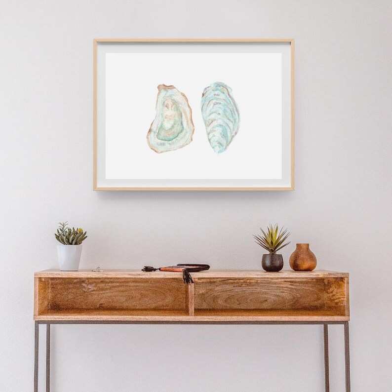 Puede incluir: Una pintura de acuarela enmarcada de dos conchas de ostra. Las conchas son de color azul verdoso claro con acentos marrones. La pintura est&aacute; sobre un fondo blanco y est&aacute; enmarcada en un marco de madera claro.