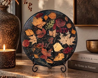 May include: A circular decorative plate featuring a dark background embedded with various pressed autumn leaves in shades of red, orange, yellow, and green. The plate is displayed upright on a black metal stand, showcasing a detailed botanical arrangement.