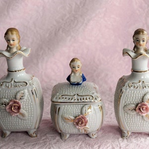 May include: A set of three white porcelain bottles with gold accents and floral designs. Each bottle features a small porcelain figurine of a woman with a blue dress and a pink rose.