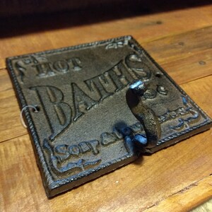 May include: A black cast iron sign with the words "Hot Baths" and "Soap and Towel" in a decorative font. The sign has a hook on the bottom.