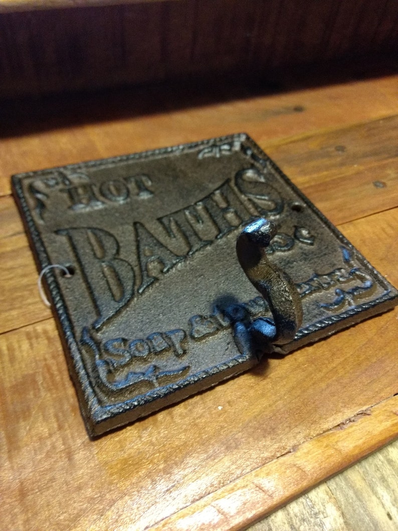 May include: A black cast iron sign with the words "Hot Baths" and "Soap and Towel" in a decorative font. The sign has a hook on the bottom.