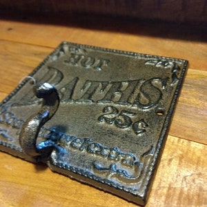 May include: A black cast iron hook with a decorative design and the words "Hot Baths 25c" on it. The hook is mounted on a wooden surface.