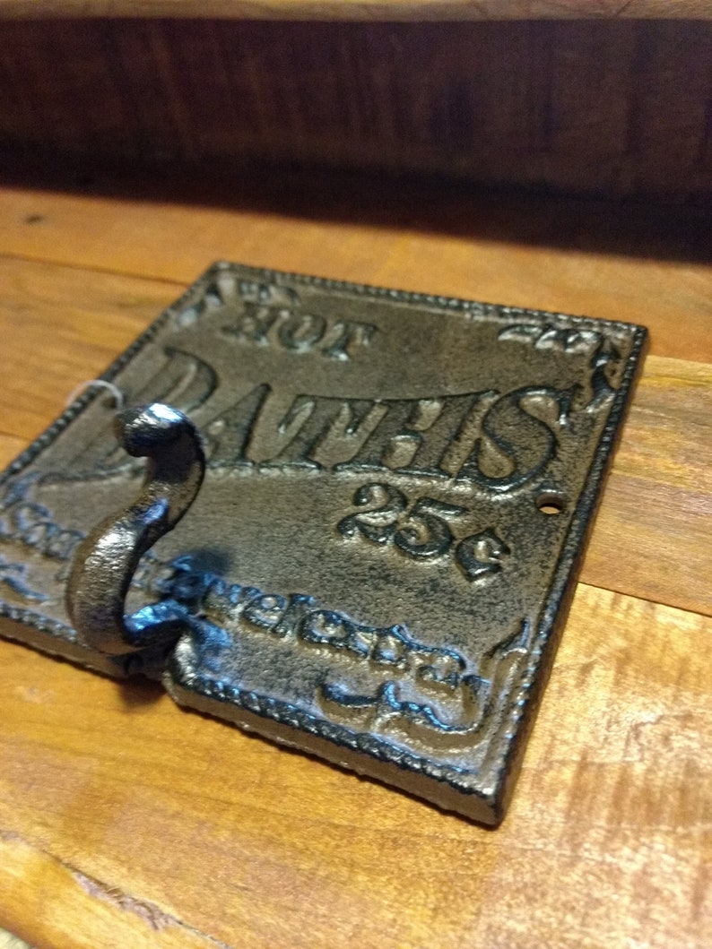 May include: A black cast iron hook with a decorative design and the words "Hot Baths 25c" on it. The hook is mounted on a wooden surface.