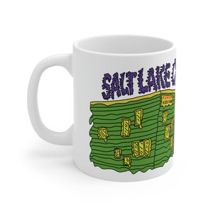 May include: White ceramic mug featuring a green building illustration and the text "SALT LAKE FUN ZONE KIDS KARAOKE" in a purple and yellow font.