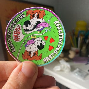 May include: A round sticker with a green background and the words "REACTIVE DOG CLUB" in a curved font. The sticker features cartoon dogs, hearts, and the text "#!&$". The sticker has a white border and is held by a hand.