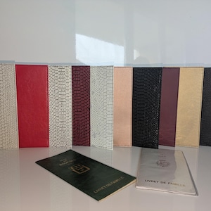 Puede incluir: Una colección de protectores de documentos de cuero sintético en varios colores, incluyendo blanco, rojo, negro y dorado. Algunos tienen un patrón de piel de serpiente. Dos folletos con texto en francés, "Livret de Famille", están delante.
