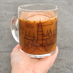 May include: A glass mug with a brown liquid inside. The mug has a black and white illustration of Hogwarts castle on the side.