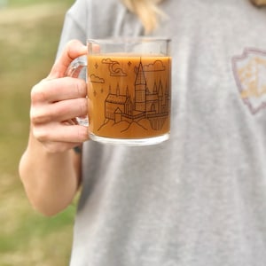 May include: A clear glass mug with a black outline of Hogwarts castle and stars. The mug is filled with a brown liquid.