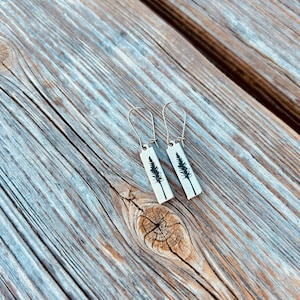 May include: A pair of rectangular earrings with a white background and a black tree silhouette design. The earrings have silver-colored ear wires and are displayed on a weathered wooden surface.