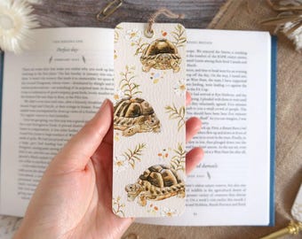 May include: A rectangular bookmark with a cream-colored background featuring a repeating watercolor pattern of brown tortoises, white flowers, and green foliage. The bookmark has a small hole at the top with a thin brown string attached.