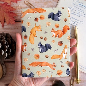 Op de afbeelding: Een notitieboek met een lichtgroene kaft met een herhalend patroon van eekhoorns, paddenstoelen en bladeren in tinten oranje, bruin en grijs. Het notitieboek wordt in een hand gehouden, met herfstbladeren en een dennenappel op de achtergrond.