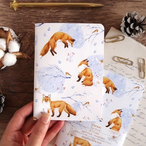 Könnte beinhalten: Ein weißes Notizbuch mit einem wiederkehrenden Aquarellmuster aus orangefarbenen Füchsen, kleinen Vögeln und blauen, verschneiten Zweigen. Der Einband zeigt den Text Carnet Notebook, 60 pages, papier recyclé, recycled paper und delphinedion.com.
