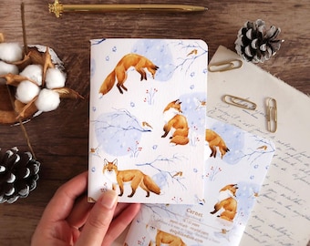 May include: Notebooks with a watercolor fox design. The cover features orange foxes, birds, and wintery branches on a white background with blue accents. A gold pen and paperclips are on a wooden surface.
