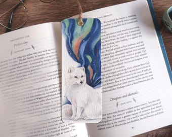 May include: A watercolor bookmark featuring a white arctic fox beneath a vibrant aurora borealis. The bookmark is rectangular with a hole at the top for a string. The bookmark is resting on an open book.