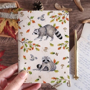 Peut inclure: Un carnet rectangulaire avec une couverture couleur crème ornée d'un motif répétitif de ratons laveurs gris, de petits oiseaux, de champignons bruns, de glands et de feuilles d'automne dans des tons de vert, orange et jaune sur papier texturé.