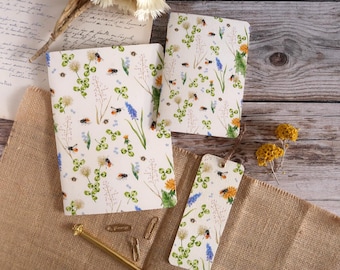 May include: A set of notebooks and a bookmark with a floral and insect pattern. The design features bees, clovers, dandelions, and blue flowers on a cream background. The items are arranged on a wooden surface with dried flowers and stationery.