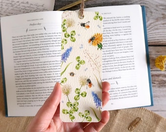 May include: A rectangular bookmark with a watercolor design featuring bees, clovers, and wildflowers. The bookmark is held open in a book. The bookmark has a hole at the top with a twine string.
