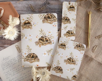 May include: A stationery set featuring a notebook, a smaller notepad, and a bookmark, all decorated with a repeating watercolor pattern of brown tortoises, green foliage, and small white flowers on a cream background, accompanied by a gold pen.