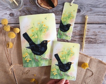 May include: A set of stationery items featuring a watercolor painting of a blackbird. The set includes a notebook, a small card, and a bookmark. The bird is depicted with yellow flowers on a light green background.