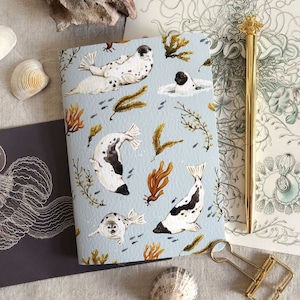 Puede incluir: Un cuaderno azul claro con una ilustración de focas, algas marinas y peces. Un bolígrafo y un clip dorados están junto al cuaderno, junto con conchas marinas y papel decorativo. El diseño del cuaderno es de estilo acuarela.