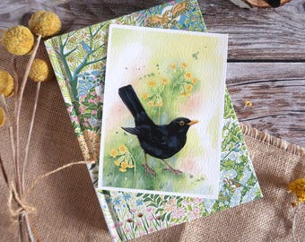 May include: A watercolor painting of a blackbird with a yellow beak and legs, set against a backdrop of yellow flowers and green foliage. The artwork is layered on patterned paper with floral and bird designs, and dried yellow flowers.