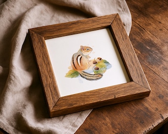 May include: Square framed watercolor art print featuring a chipmunk with brown and tan fur, sitting near acorns and green leaves. The artwork is set against a white background and framed in a dark brown wooden frame.