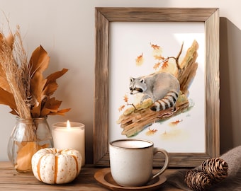 May include: A framed watercolor print of a raccoon on a log, with fall leaves. The print is displayed on a wooden surface with a lit candle, a pumpkin, a coffee mug, and decorative dried foliage in a glass vase. Two pinecones are also present.