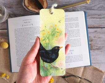 May include: A hand-painted bookmark featuring a blackbird with an orange beak, set against a backdrop of yellow flowers and green foliage. The bookmark has a cream-colored string and is held in front of an open book.