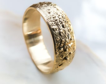 May include: A gold ring with a textured surface. The band features a series of small, irregular bumps and indentations, giving it a unique, organic appearance. The ring is set against a white background.