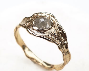 May include: A gold ring with a textured band and a central, gray, faceted gemstone. The ring's design has an organic, flowing shape, with a slightly irregular band. The gemstone is set in a bezel setting, and the ring is displayed against a white background.