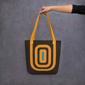May include: A brown tote bag with yellow straps and a geometric design featuring concentric circles in orange and brown with a blue line in the centre.