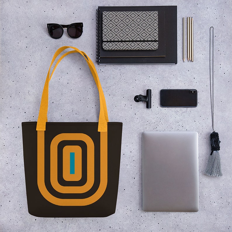 May include: A brown tote bag with yellow straps and a geometric design in orange and teal. The design features concentric circles with a vertical line in the centre.