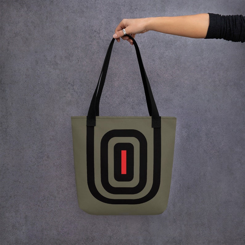 May include: A green tote bag with black straps and a black and red geometric design. The design features concentric circles with a red vertical line in the center.