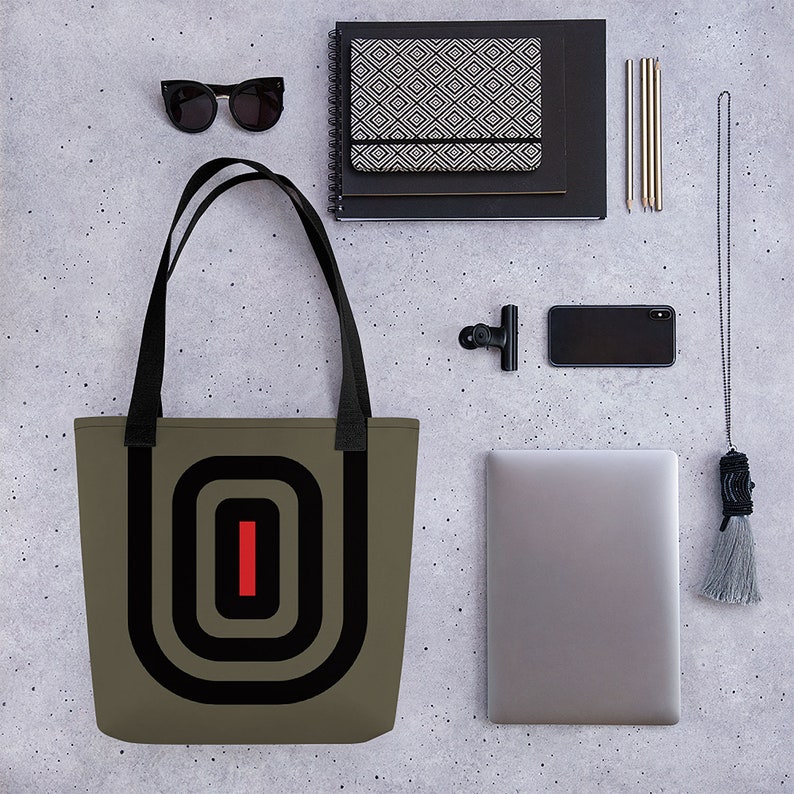 May include: A khaki tote bag with black straps and a black and red geometric design. The design features concentric circles with a red vertical line in the center.