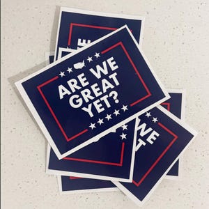 May include: A stack of blue and red stickers with white text that reads "ARE WE GREAT YET?" with stars and a small image of the United States of America.