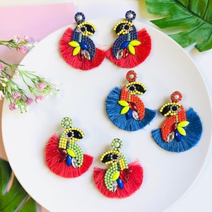 Op de afbeelding: Een set van zes kleurrijke kralen oorbellen met franjes. De oorbellen zijn in verschillende kleuren, waaronder rood, blauw, groen, geel en zwart. De franjes zijn gemaakt van rood, blauw en groen garen.