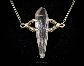 May include: A clear quartz crystal pendant necklace. The crystal is held by two silver-toned claw charms, suspended from a delicate chain. The necklace is set against a black background, highlighting the crystal's clarity and the intricate details of the claws.